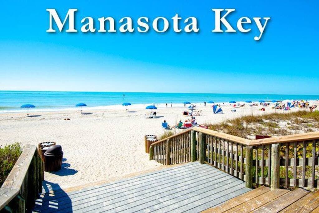 A sandy beach stretches towards the horizon, with colorful umbrellas dotting the landscape. The calm blue waters of the Gulf of Mexico contrast against a clear sky. A wooden deck leads from the foreground to the beach, offering easy access to the coastal paradise.