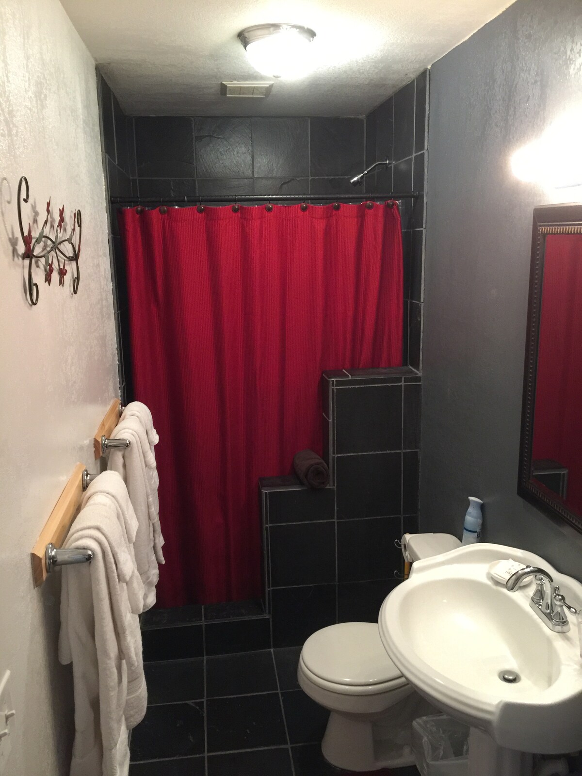 A compact bathroom features black tiled walls and floor, with a red shower curtain adding a pop of color. A white sink with a mirror above is complemented by neatly hung towels on a wooden rack. A small soap dispenser and trash bin complete the space.
