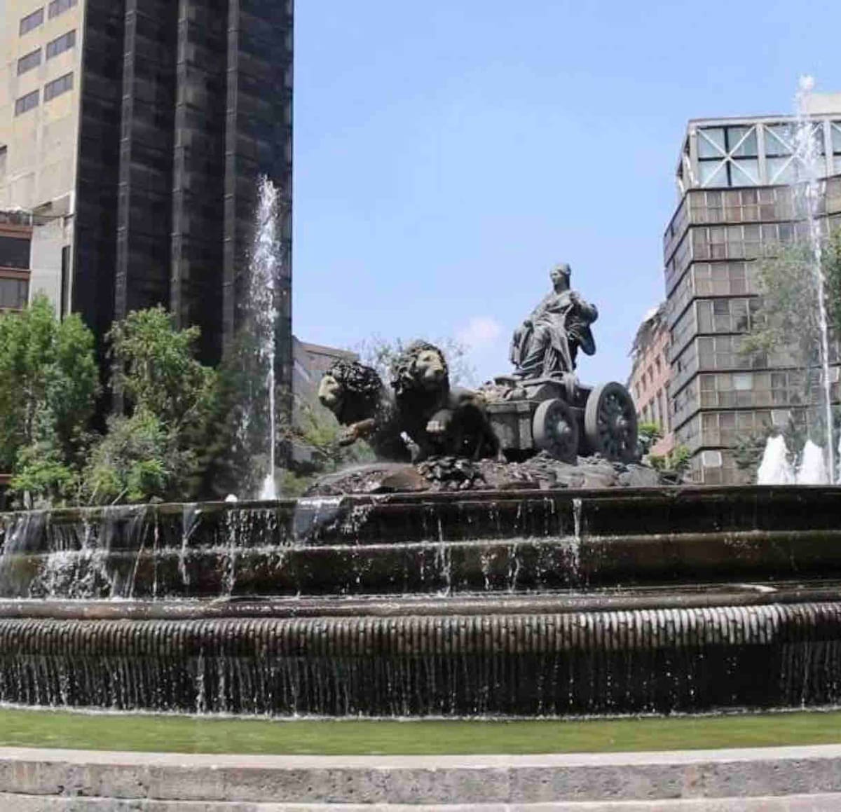 A fountain featuring intricate sculptures, including a figure on a chariot drawn by lions. Water cascades down in multiple streams, creating a serene environment. Surrounding greenery enhances the setting, with modern buildings visible in the background.