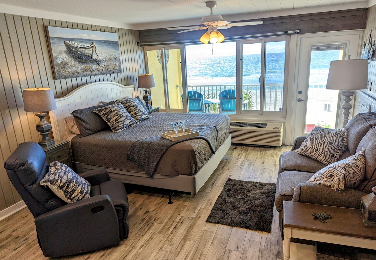 A spacious living area includes a king bed adorned with decorative pillows, paired with a cozy seating arrangement featuring two recliners and a sofa. Large windows provide natural light and stunning views, while a small area table sits ready for refreshments.