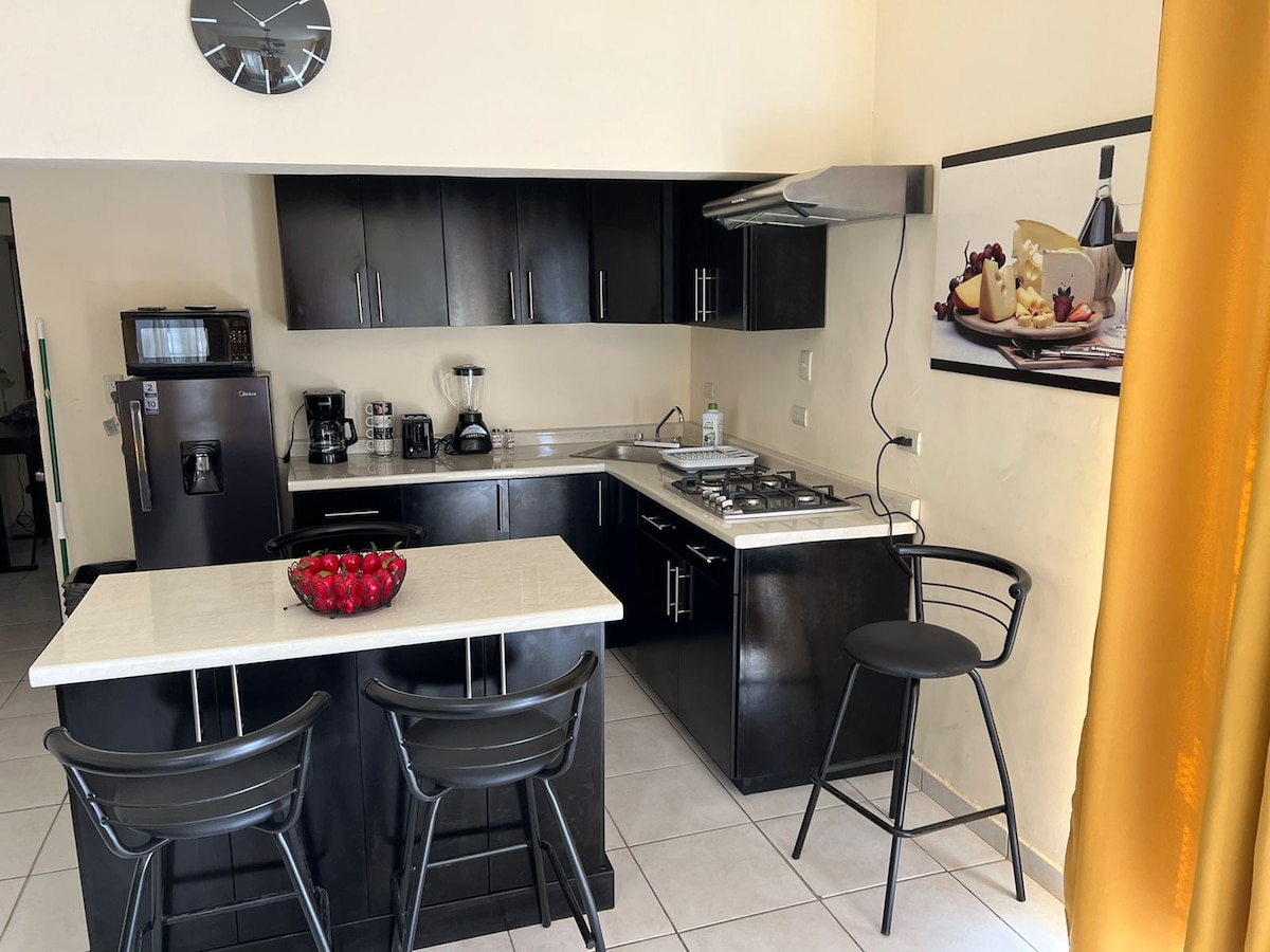 A modern kitchen features sleek black cabinetry and a light-colored countertop. Bar stools are positioned at a compact island, while various appliances, including a coffee maker and a blender, are visible. A framed piece of art depicting fruit adds a touch of color to the space.