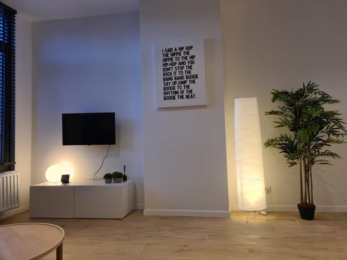A cozy living area is presented with a mounted television and a minimalist white cabinet. A rounded lamp emits soft light beside a tall, decorative plant. A large wall art piece features text in a bold font, adding a contemporary touch to the space.