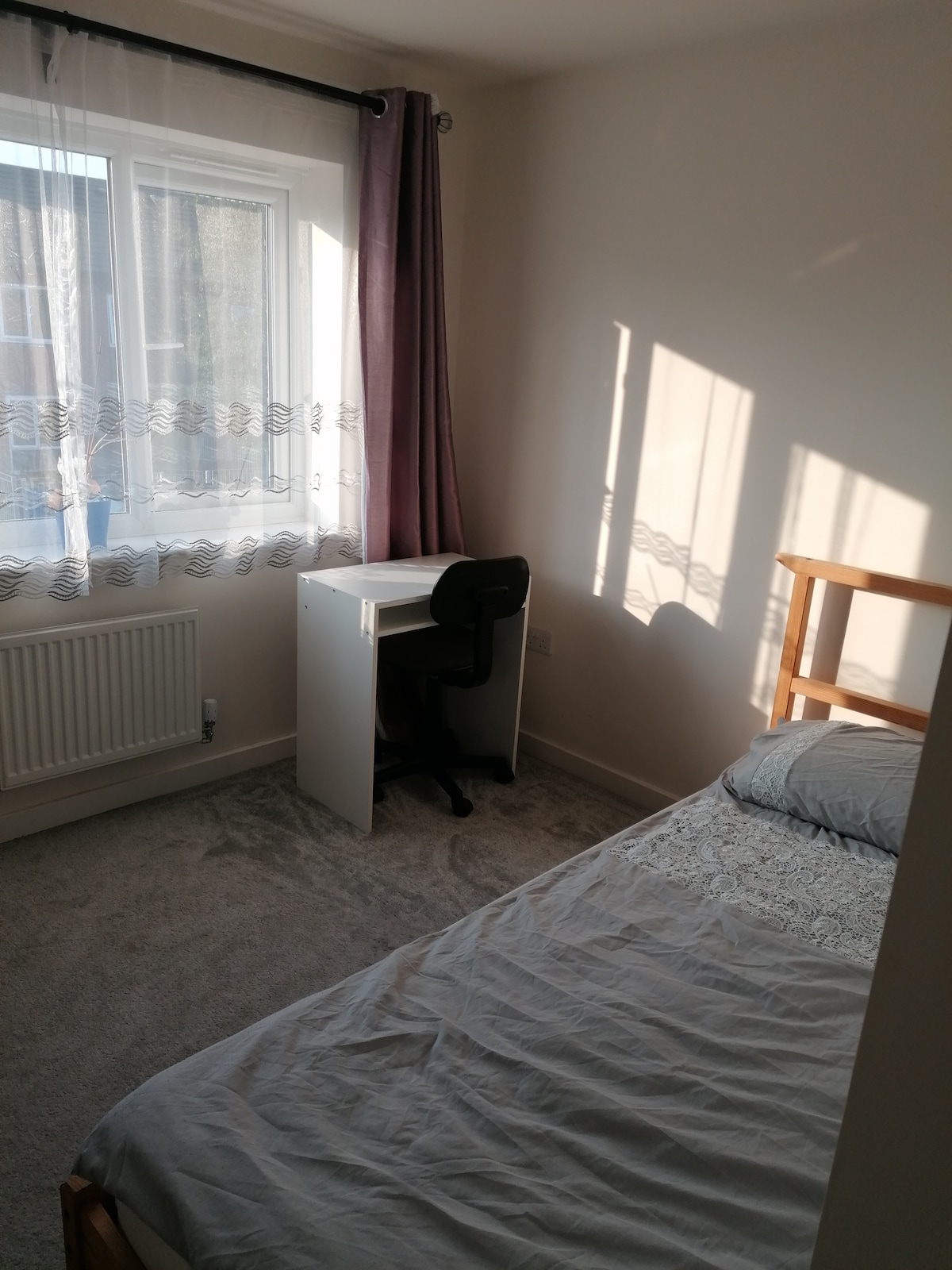 A well-lit room features a single bed with soft bedding positioned beside a spacious window. Natural light filters through sheer curtains, creating a bright ambiance. A small desk with a chair is neatly arranged against the wall, offering a functional space for work or leisure.