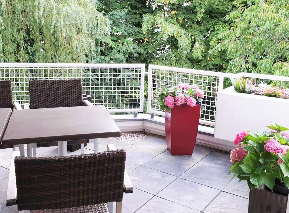 A balcony space features a dark wicker dining table accompanied by four matching chairs. Colorful pots with blooming flowers, including pink hydrangeas, add a touch of nature. The backdrop consists of lush green trees, providing a sense of privacy and tranquility.