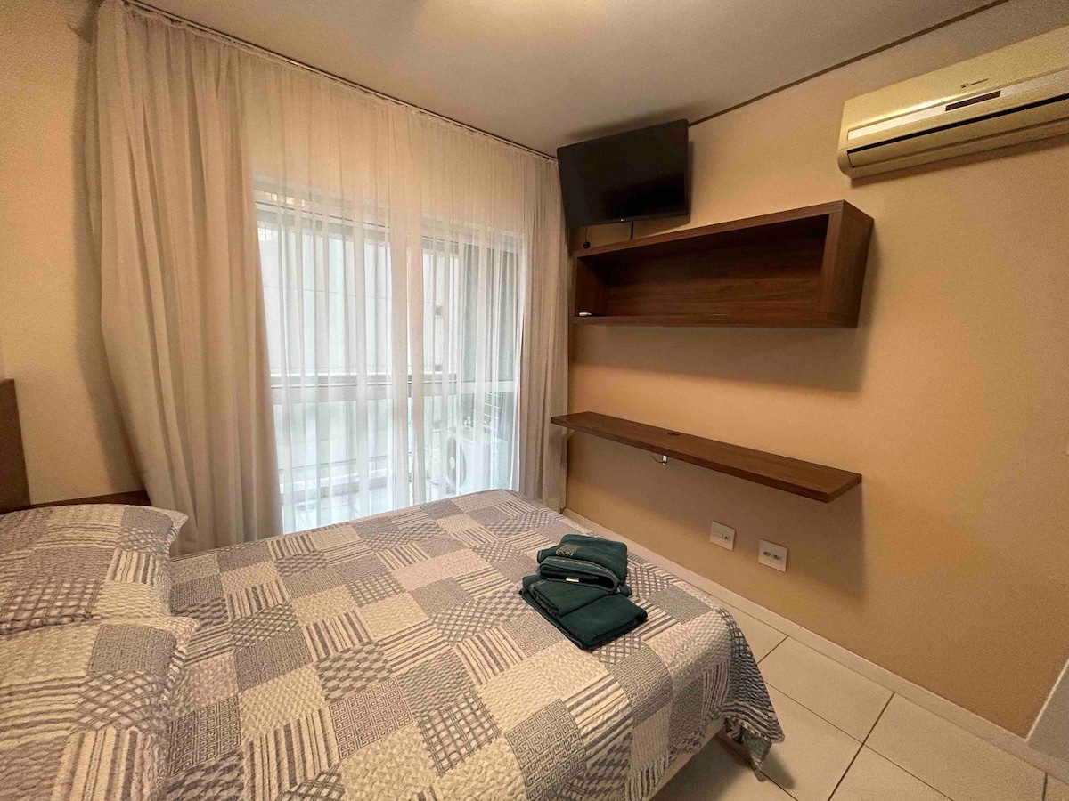 A comfortable sleeping area features a spacious bed with a patterned quilt and neatly arranged towels. A wooden shelf and a mounted flat-screen TV are positioned on the wall, while natural light enters through sheer curtains covering the window.