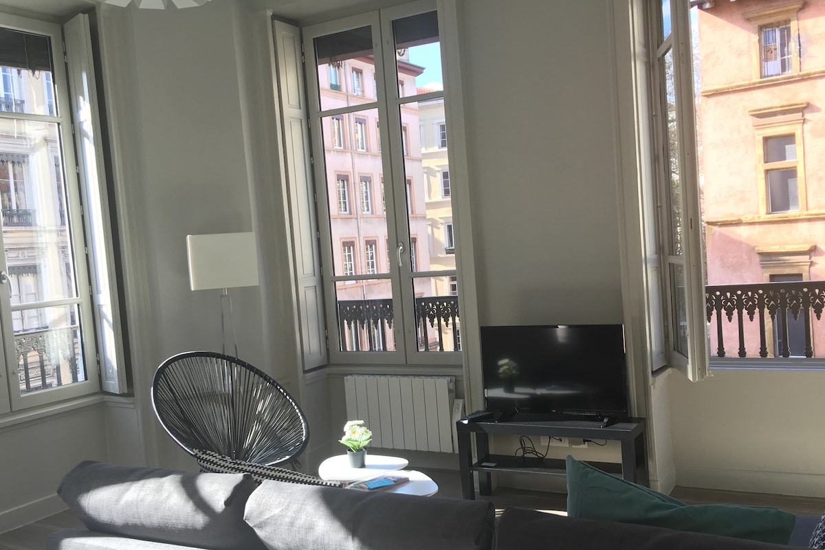 A bright living area is illuminated by natural light streaming through large windows. Modern furniture includes a black accent chair and a television on a simple stand. The views showcase the charming architecture of the surrounding buildings.
