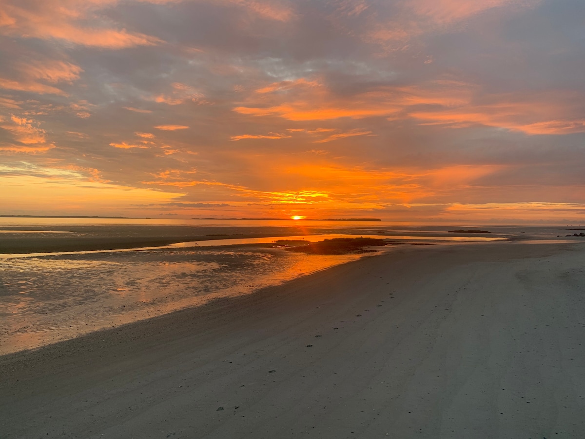 A serene beach scene at sunset, where the sky is filled with hues of orange and gold. The calm water reflects the vibrant sky, and footprints are visible in the fine sand leading along the shoreline.
