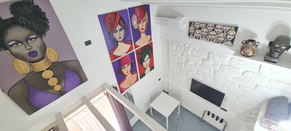 A ceiling view captures the stylish interior of the studio, featuring a variety of artistic wall displays. A compact dining table is positioned near a wall-mounted television, while a textured stone surface adds visual interest. Air conditioning is visible, enhancing comfort within the space.