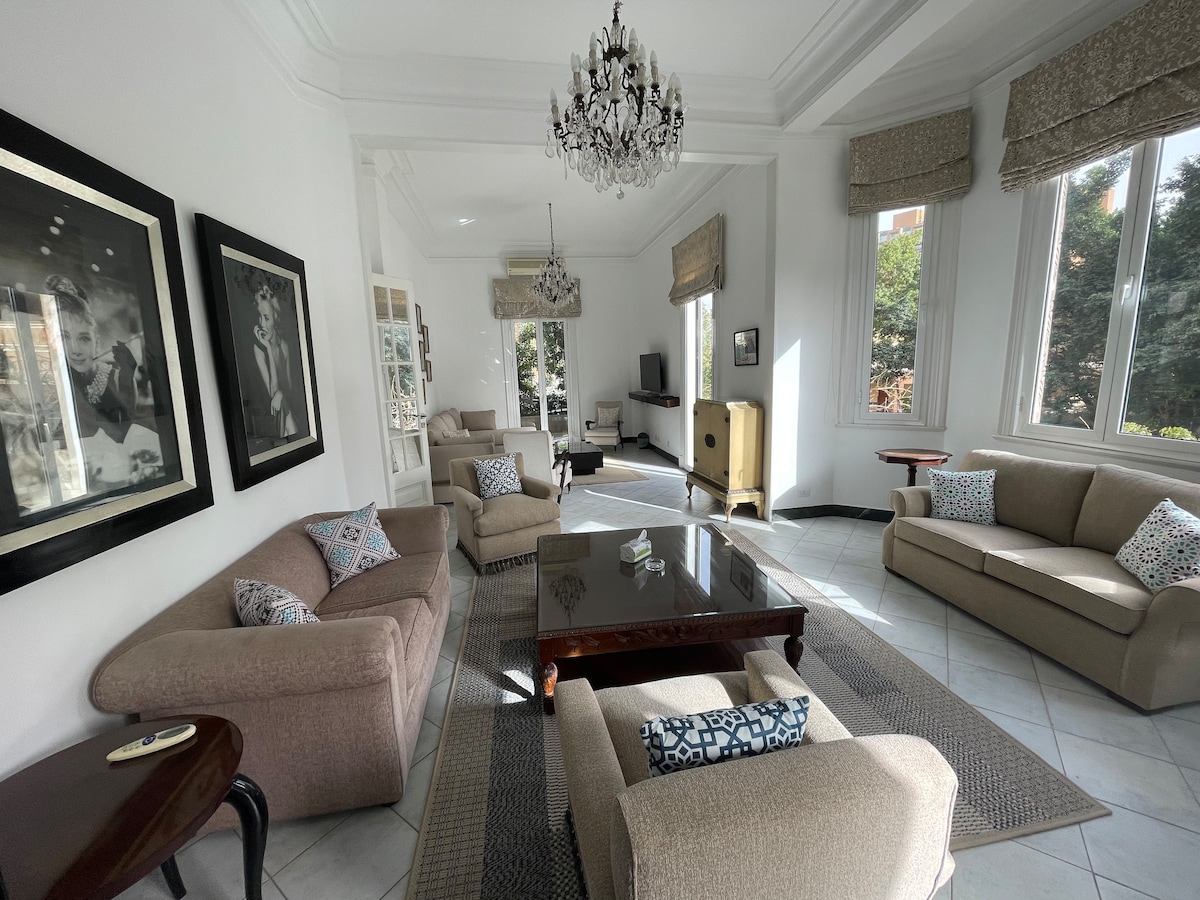 The spacious living area features two sets of comfortable sofas arranged around a polished wooden coffee table. Large windows allow natural light to fill the room, with soft drapes partially covering them. Decorative artwork and a chandelier contribute to the elegant ambiance.