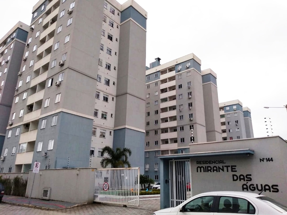 The apartment building is shown from the exterior, featuring a modern design with a light gray facade. Notable elements include a gated entrance with the name 'Residencial Mirante das Águas' displayed prominently. Multiple floors and windows are visible, contributing to a spacious impression.