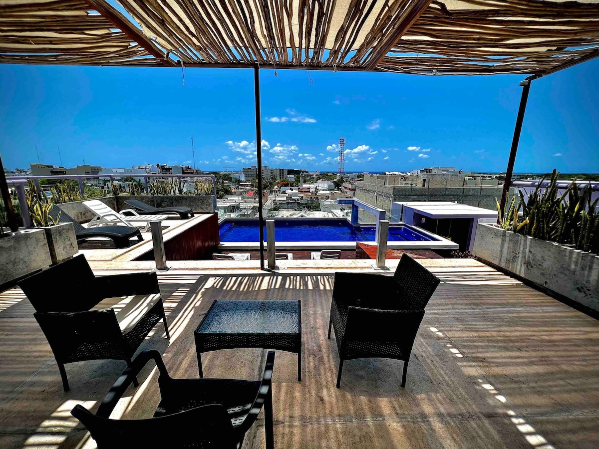 An outdoor seating area features black wicker chairs and a small table, positioned beneath a shaded canopy. A swimming pool is visible in the background, with panoramic views of the surrounding cityscape and clear blue sky.