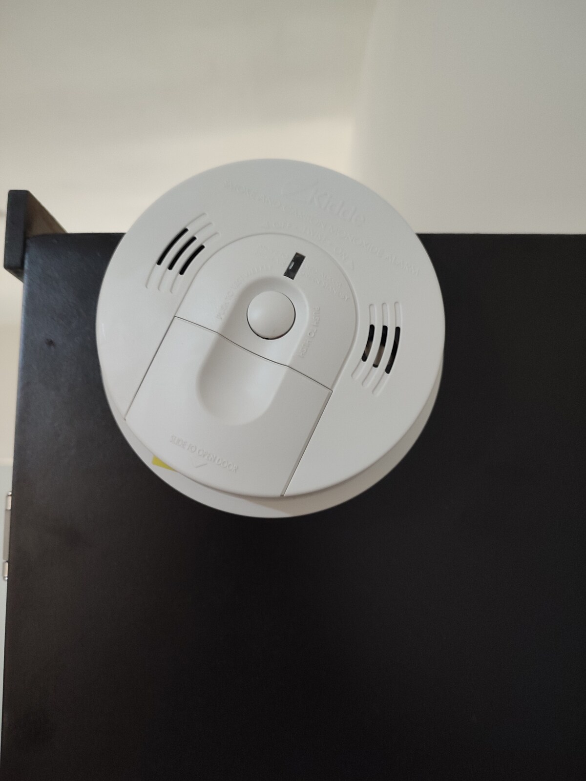 A smoke detector is mounted on a dark surface, featuring a central button and multiple ventilation slots. The design is simple and functional, emphasizing safety within the home.