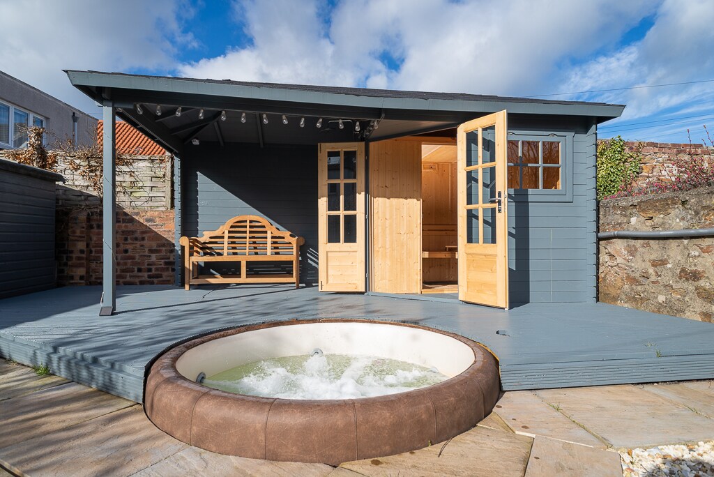 An outdoor area features a round hot tub, surrounded by a wooden deck. Nearby, a wooden bench sits under a covered porch with double doors, allowing entry to a sauna. The setting includes a mix of stone and wooden elements, under a partly cloudy sky.