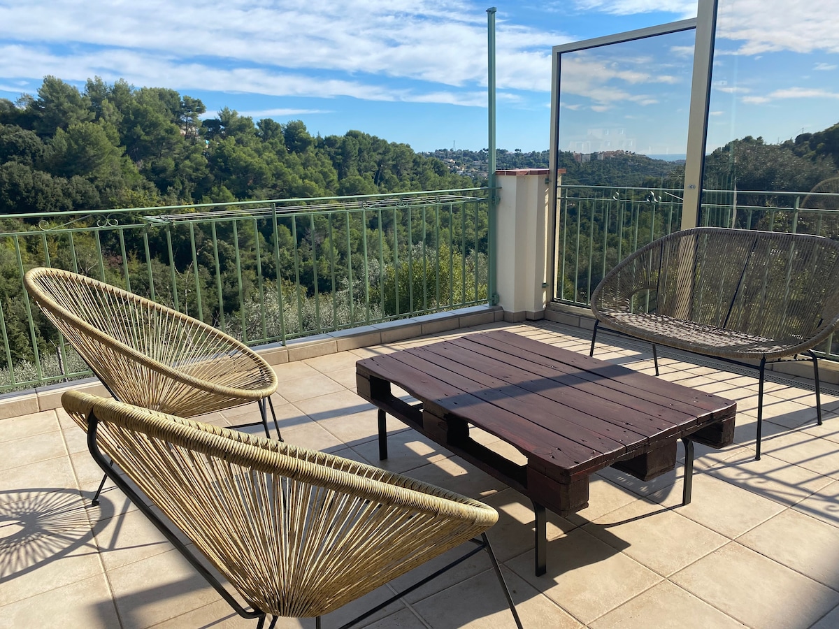 A spacious terrace features two woven chairs and a low wooden table, set against a backdrop of lush green hills under a clear blue sky. The railing provides an unobstructed view of the surrounding nature.