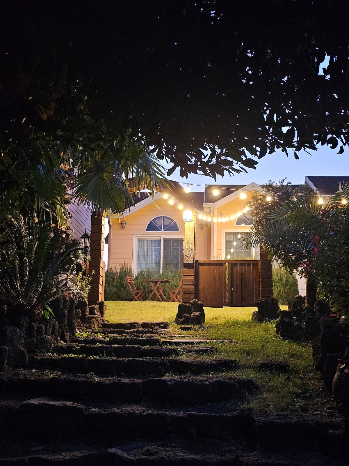 A pathway leads through lush greenery towards an entrance adorned with warm string lights. The inviting exterior showcases a wooden door and a small seating area with two chairs. Soft illumination highlights the charm of the surrounding foliage, creating a welcoming ambiance.