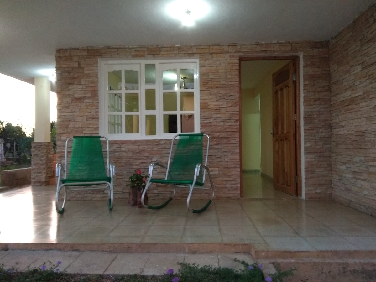 Two green rocking chairs are positioned on a tiled porch in front of a stone wall. A door leads into the interior, and a window allows natural light to filter in, enhancing the space's openness. Potted plants add greenery to the welcoming entrance.
