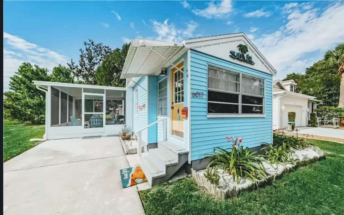 A charming blue house is framed by lush green grass and vibrant landscaping. The entrance features a small flight of steps leading to a covered porch. A screened-in patio is visible to the left, providing an inviting outdoor space filled with natural light.