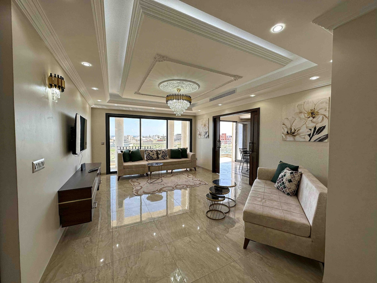 A spacious living room features elegant lighting fixtures, with a chandelier suspended above. Comfortable seating arrangements are positioned along the walls, complemented by a modern coffee table. Large windows allow natural light to illuminate the marble flooring, enhancing the open atmosphere.
