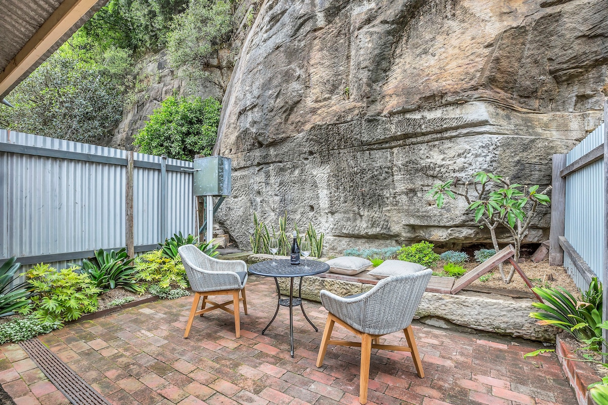 An outdoor entertainment area is showcased, featuring a round table and two armchairs positioned on a brick patio. Lush greenery surrounds the space, with a large rock face providing a natural backdrop.