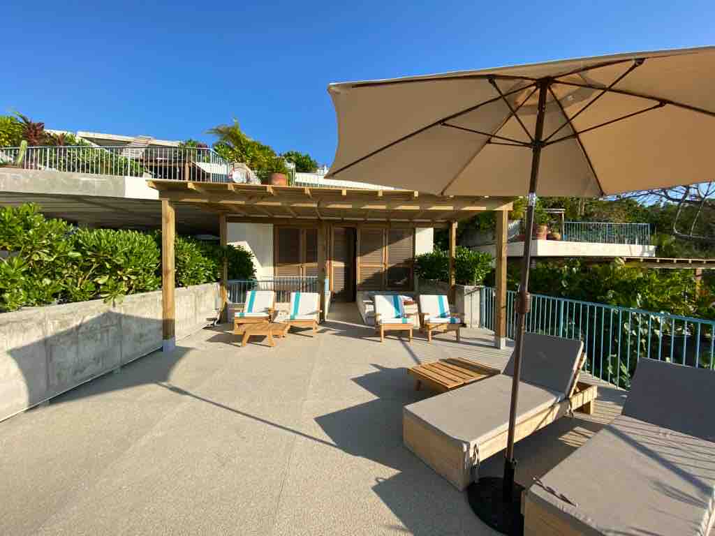 A spacious outdoor terrace is equipped with lounge chairs and a shaded area under a large umbrella. Greenery lines the perimeter, providing a natural backdrop. Glass doors lead into the interior space, inviting relaxation in a bright, sunlit environment.