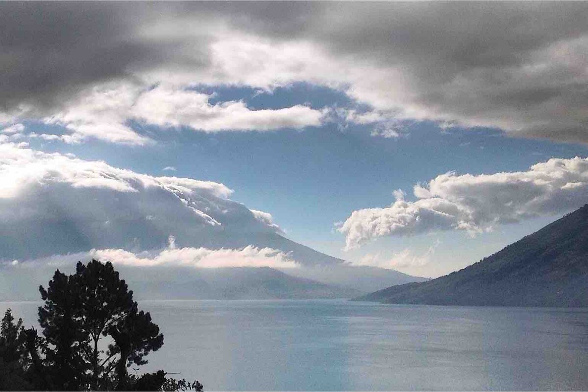 A serene view of Lake Atitlán unfolds under a cloudy sky, with mountains framing the horizon. Soft sunlight filters through the clouds, illuminating the lake's surface, which reflects the colors of the sky. Lush trees are visible in the foreground.