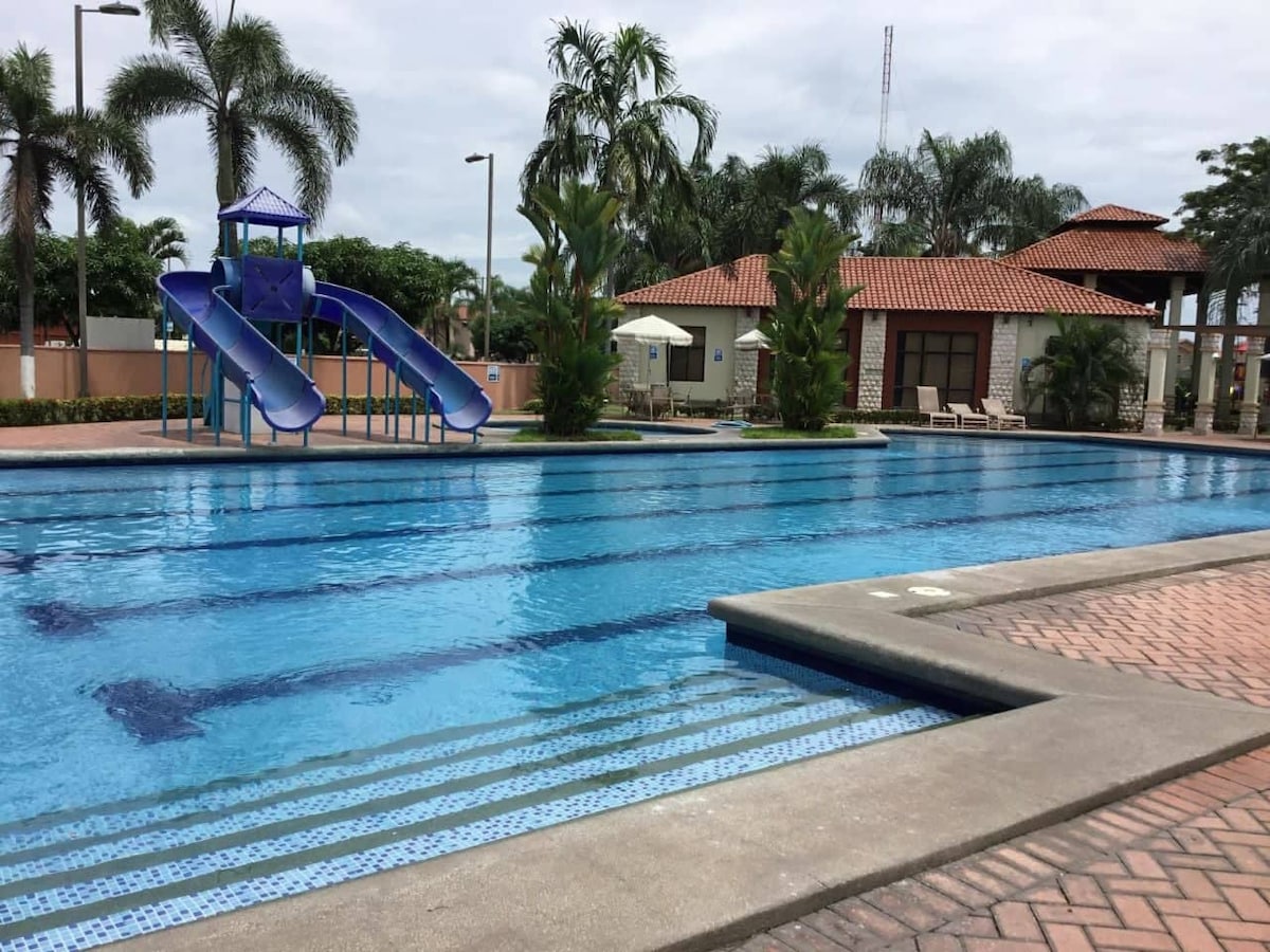 A large swimming pool is featured, accompanied by a blue water slide. Lush palm trees and well-maintained landscaping surround the pool area. A shaded seating area and a building with a terracotta roof are visible in the background, providing additional amenities for visitors.
