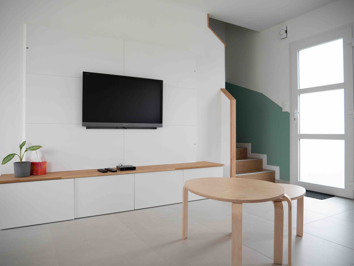 A bright living area is equipped with a wall-mounted television above a sleek white entertainment unit. A light wood coffee table is positioned in the center, complemented by a small potted plant. A staircase leads to the upper floor, enhancing the open space.