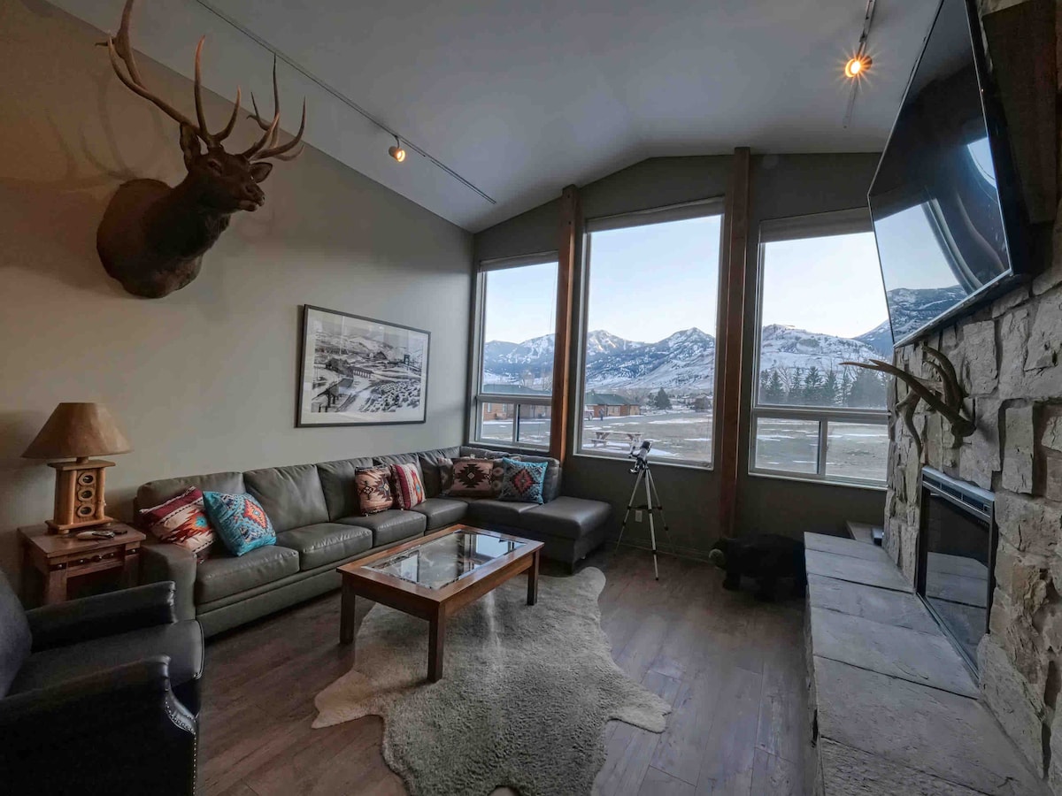 A spacious living room is showcased with large windows providing views of snow-capped mountains. A mounted elk head and artwork decorate the walls. Comfortable seating is arranged around a coffee table on a soft area rug, with a stone fireplace and a television enhancing the cozy setting.