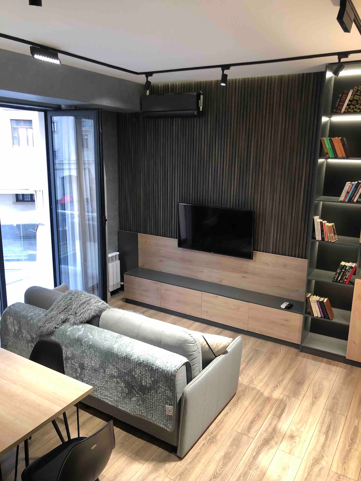A modern living area is highlighted by a flat-screen TV mounted on a wooden panel. A comfortable gray sofa and a wooden dining table are visible, with shelves showcasing books along the wall. Large windows allow natural light to enter the space.