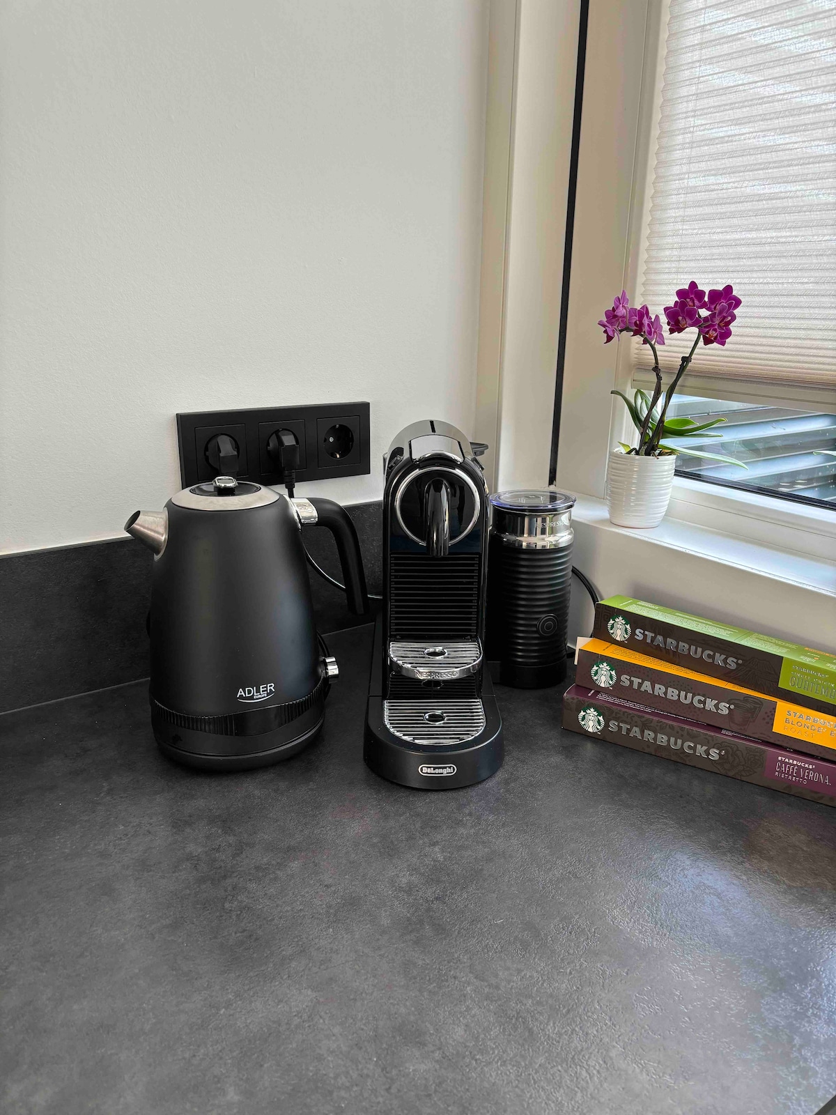 A modern kitchen counter is featured, displaying a black kettle, a coffee machine, and a travel mug. A small white pot with an orchid adds a touch of greenery. Several boxes of Starbucks coffee are stacked nearby, contributing to a well-organized and functional space.