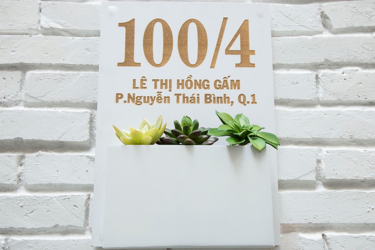 A clean white wall features a decorative sign with the address '100/4 Lê Thị Hồng Gấm, P. Nguyễn Thái Bình, Q.1'. Above the address, a small planter holds various green succulents, adding a touch of natural texture to the design.