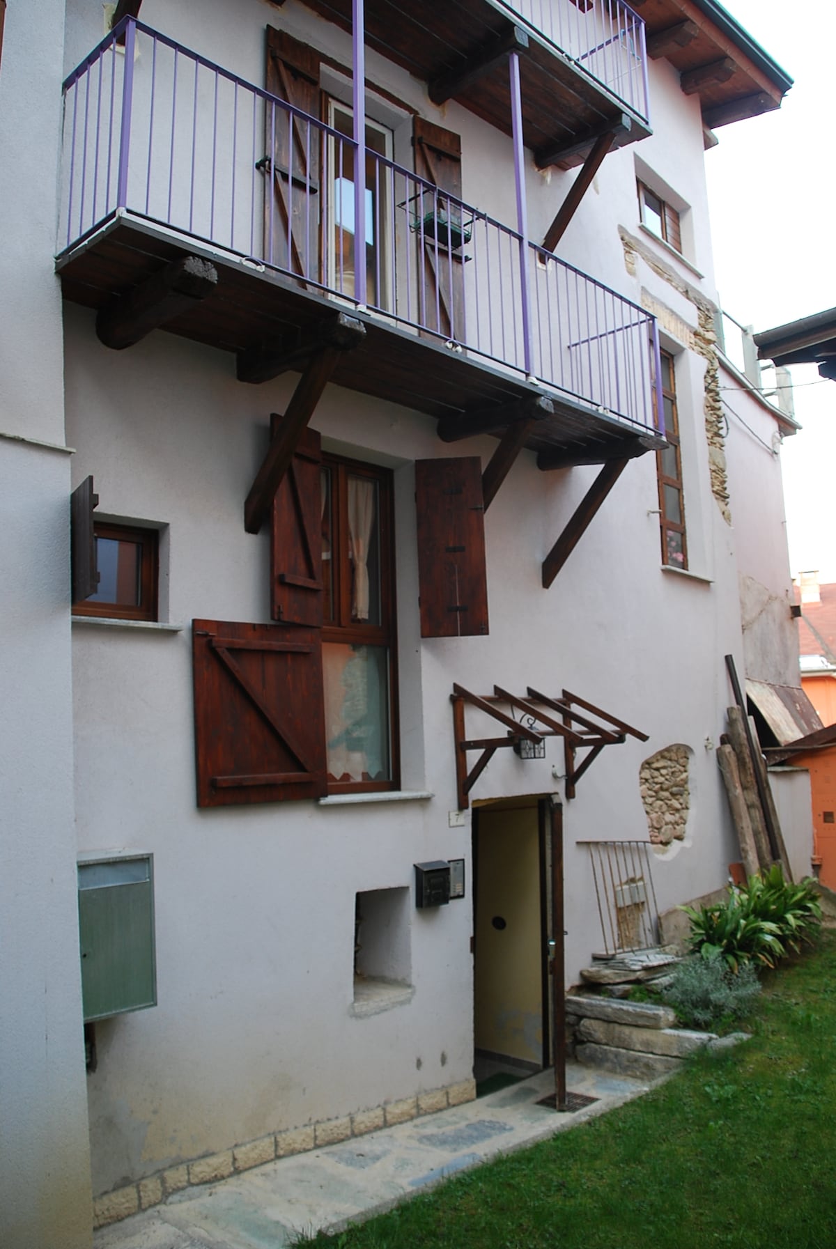 An exterior view of a multi-story building is shown, featuring a balcony supported by wooden beams. Closed wooden shutters cover the windows. A small door with a simple awning leads to the entrance, with a staircase leading up to the balcony area. Lush greenery is visible along the base.
