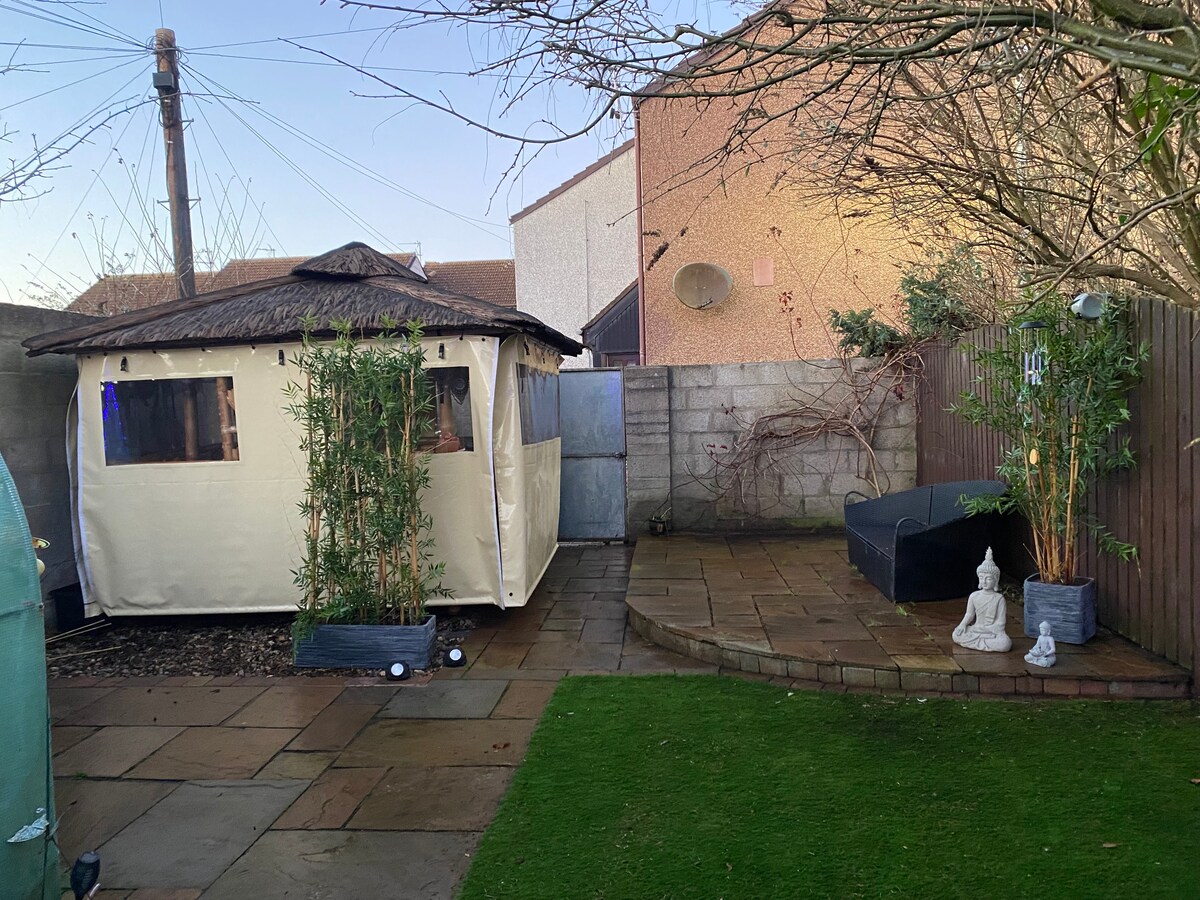 An outdoor area features a spacious paved patio surrounded by lush greenery and bamboo plants. A heated gazebo is visible, offering a shaded retreat, while a stylish seating area provides a comfortable spot for relaxation. Decorative elements like statues add character to the garden.