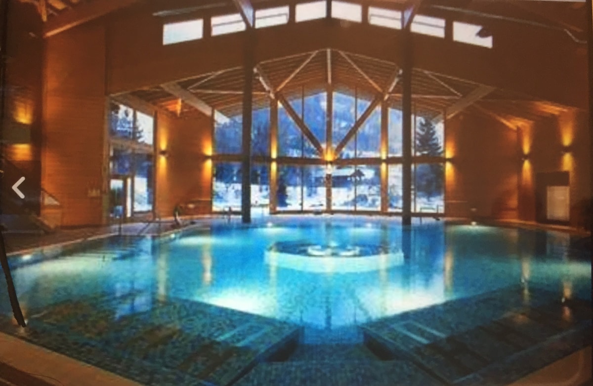 An indoor swimming pool is surrounded by large windows, allowing natural light to illuminate the space. The pool features a circular center and step entry options. Wooden beams and warm-toned walls contribute to a serene environment, with views of the surrounding landscape visible through the windows.