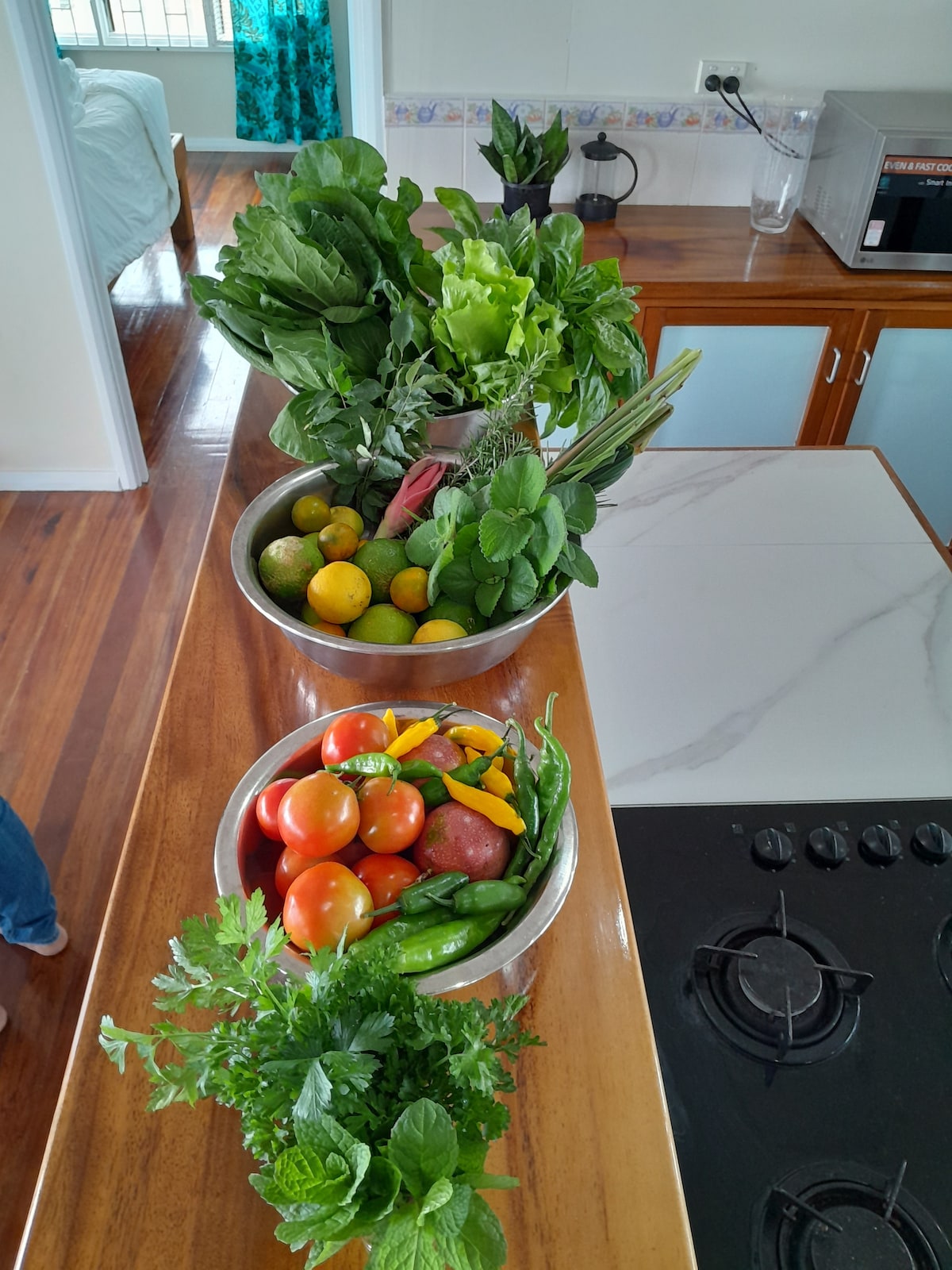 Two large bowls filled with fresh produce are positioned on a kitchen countertop. One bowl contains green leafy vegetables, citrus fruits, and herbs, while the other holds tomatoes, apples, and green chili peppers. A modern stovetop and part of the kitchen layout are also visible.