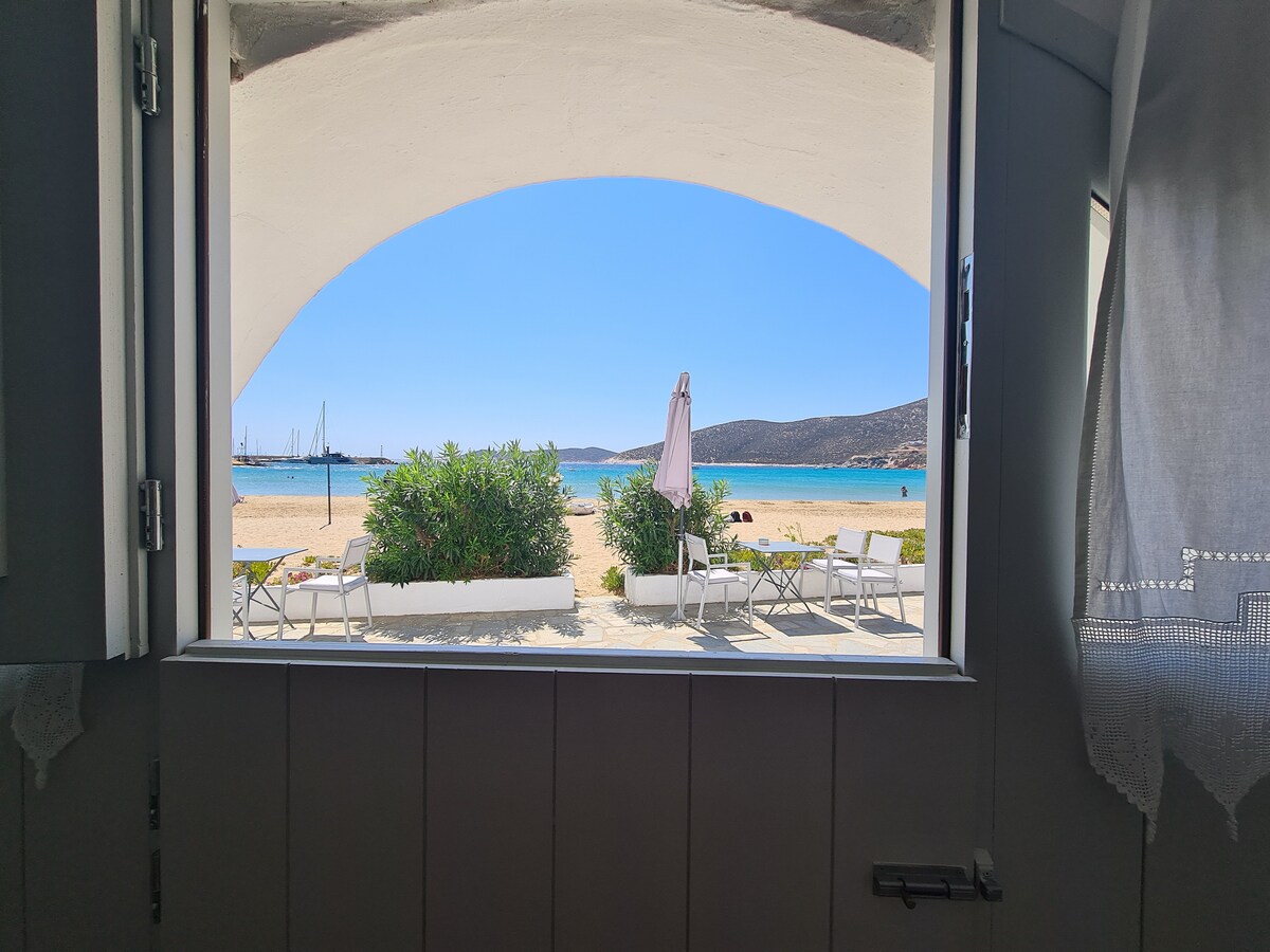 A scenic view is framed by an archway, showcasing a sandy beach lined with lounge chairs and umbrellas. The tranquil blue sea is visible along with distant hills, creating a serene coastal atmosphere. Natural light from the outside illuminates the room.