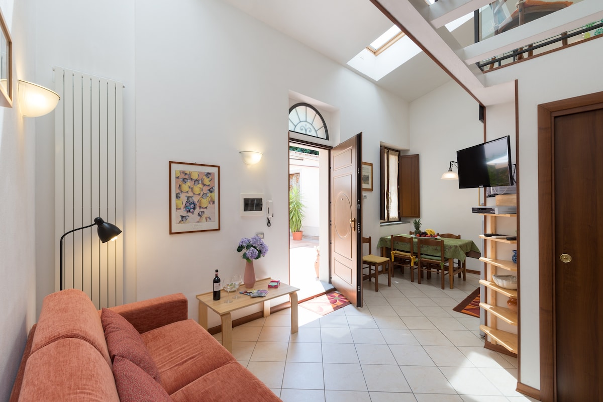 A bright living area features a cozy orange sofa and a wooden coffee table. An entrance door opens to a charming courtyard, while a television is mounted on the wall. Skylights above enhance the spacious ambiance, complemented by a dining area set with a table and chairs.
