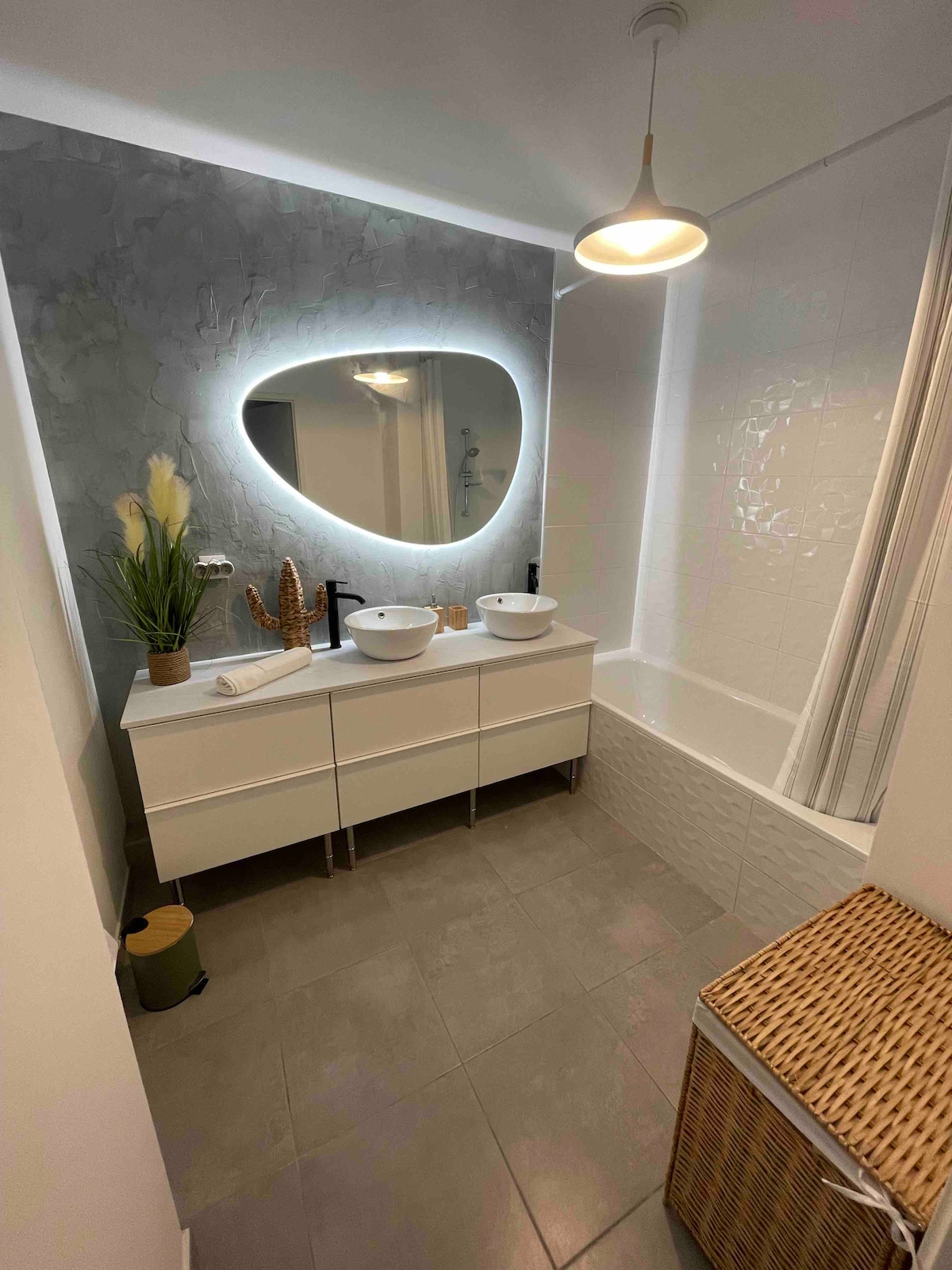 A modern bathroom is showcased, featuring a double vanity with two circular basins under a uniquely shaped illuminated mirror. A bathtub is positioned beside the vanities, and a wicker basket is discreetly placed for storage. Soft lighting enhances the clean, neutral tones throughout.