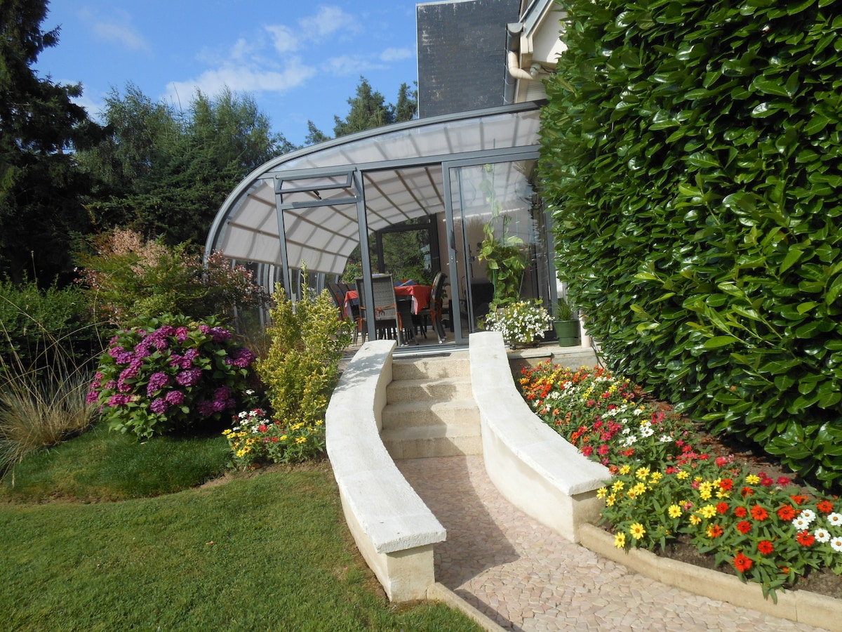 A bright and airy veranda is framed by lush greenery and colorful flowering plants. A stone pathway leads to the entrance, inviting guests to enjoy the surrounding beauty. The structure features large glass panels, enhancing the connection with the outdoors.