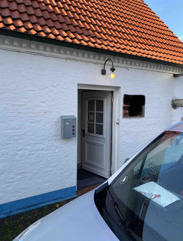 The exterior entrance features a white wall with blue trim and a welcoming light above the door. A mail slot is visible, and a vehicle is parked nearby, indicating the convenience of access. The red-tiled roof adds character to the building's design.