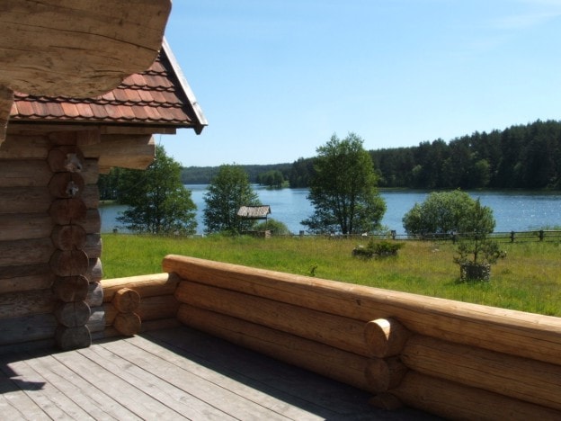 A wooden deck overlooks a tranquil lake, framed by lush greenery and distant trees. The clear blue sky reflects on the water's surface, creating a serene outdoor atmosphere. Log railings add an authentic touch to the rustic setting.