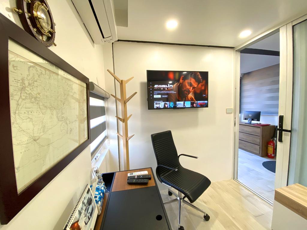 A workspace is featured in the image, with a desk, black chair, and a wall-mounted television displaying content. A map is framed on the wall, while a coat rack stands nearby. Natural light is introduced through a glass door leading to an outdoor space.