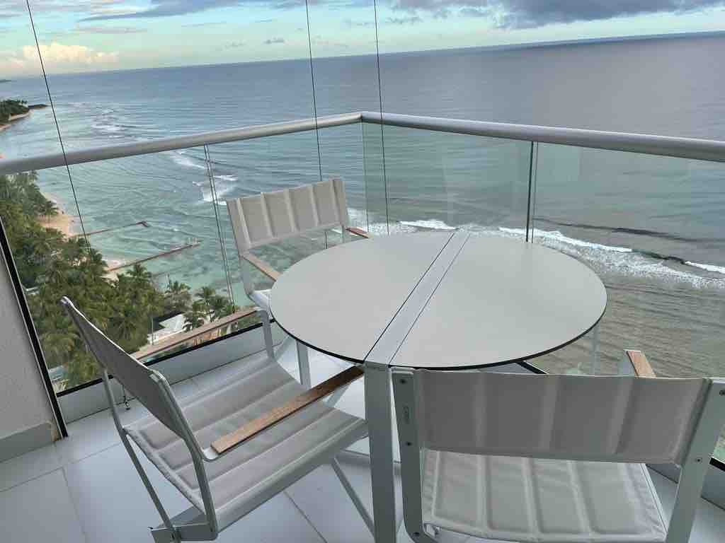 A balcony seating area is visible, featuring a round glass table surrounded by two white chairs. The ocean view extends to the horizon, with gentle waves lapping at the shore, framed by palm trees and a sandy beach.