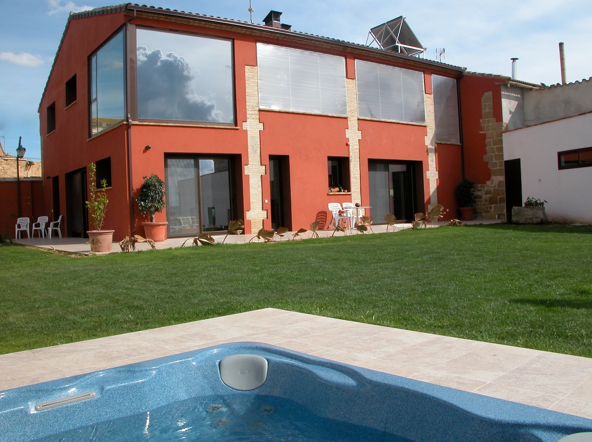 A spacious garden area showcases a jacuzzi in the foreground, with a modern two-story building in the background. Large glass doors provide a view of the interior, while patio chairs are placed underneath the overhangs. Lush green grass surrounds the structure, enhancing the outdoor space.