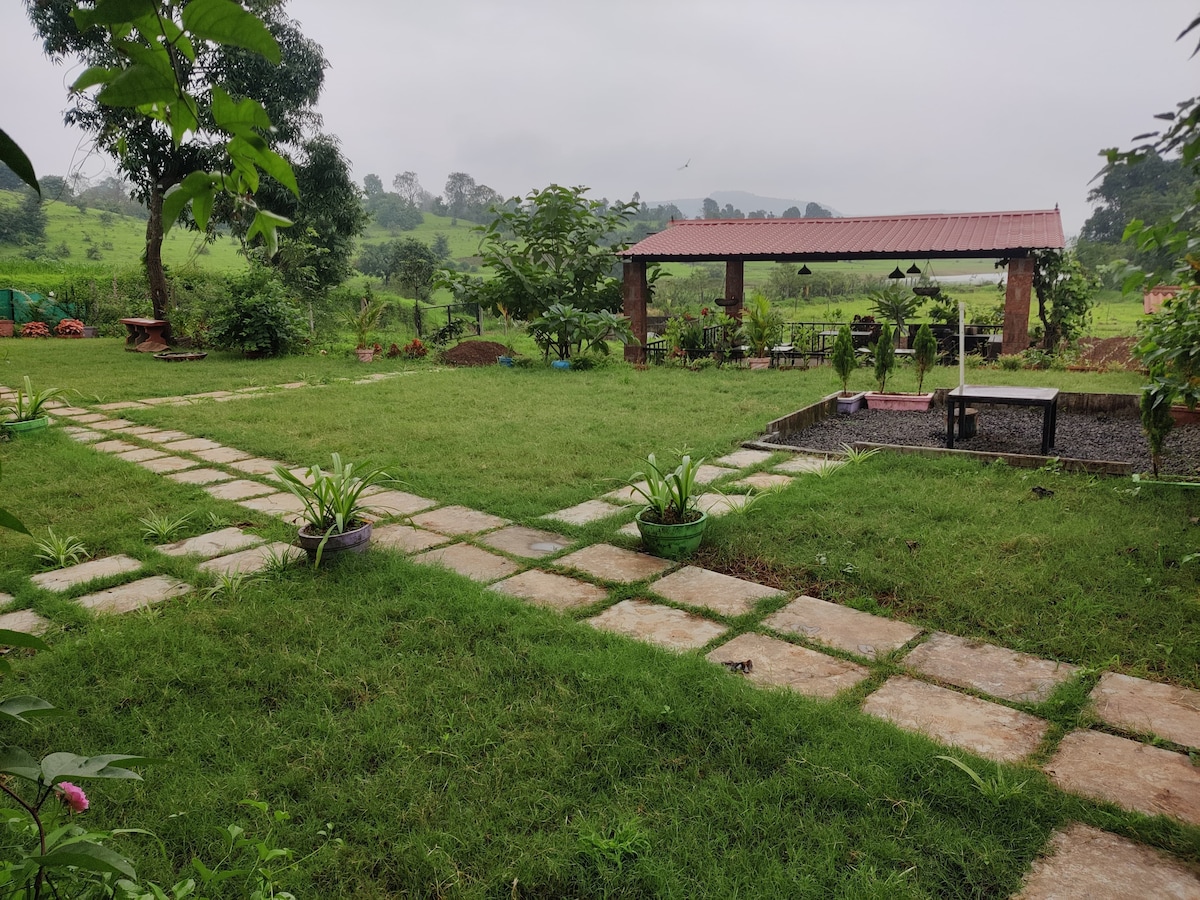 A lush green garden is featured with a stone pathway leading through the grass. Low plant beds and picnic areas are visible, along with a sheltered seating space adorned with hanging lights, surrounded by hills and trees in the background under a cloudy sky.