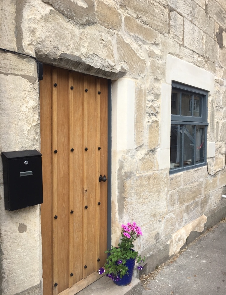 The exterior of the cottage features a rustic wooden door with decorative metal studs, framed by Cotswold stone walls. A window with dark trim complements the natural stone. A blue flower pot with blooming pink flowers adds a touch of color at the entrance.