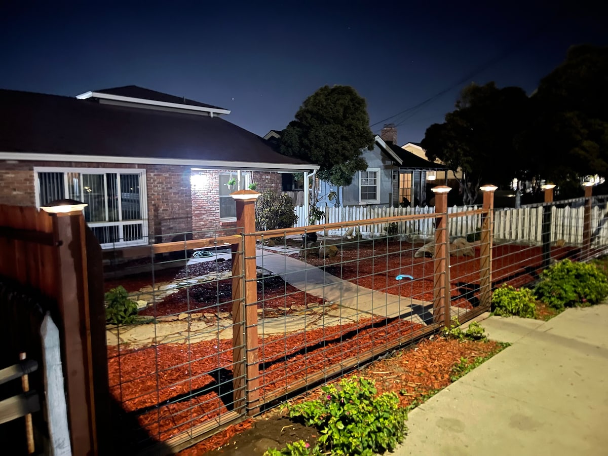 The exterior view of the inviting brick house is captured at night, featuring soft lighting illuminating the pathway. A wooden fence with metal accents encloses the landscaped front yard, where red mulch adds a touch of color, and well-maintained shrubs line the walkway.