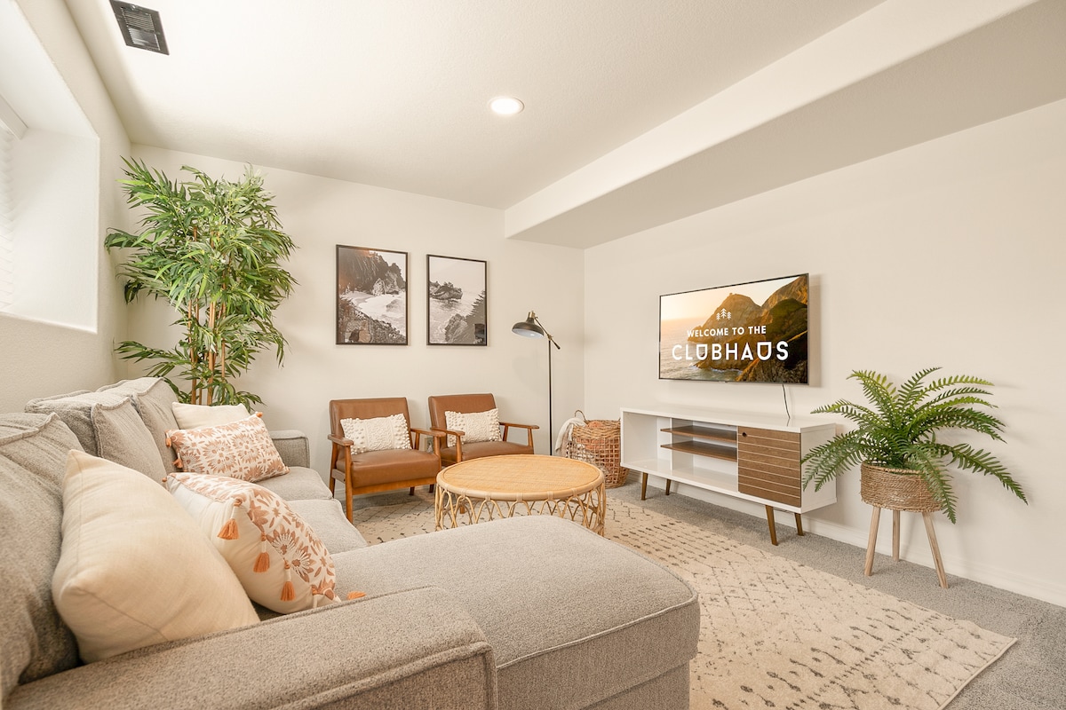 A cozy living area features a light-colored sectional sofa adorned with decorative pillows. Two wooden chairs provide additional seating. A circular coffee table sits in the center, complemented by greenery and wall art. A large TV is mounted on the wall, enhancing the space.