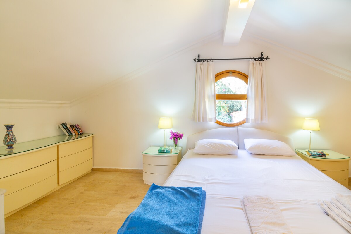 A bright bedroom features a spacious bed with crisp white linens, flanked by two bedside lamps. Light filters through a round window, illuminating light-colored furnishings, including a dresser and shelves with books. A touch of color is added by a small flower arrangement.