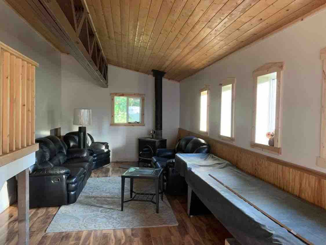 A spacious living area is highlighted by a wooden ceiling and natural light from multiple windows. Comfortable black leather seating accommodates guests, complemented by a small coffee table at the center. A sleek wood stove adds warmth to the space, enhancing relaxation.